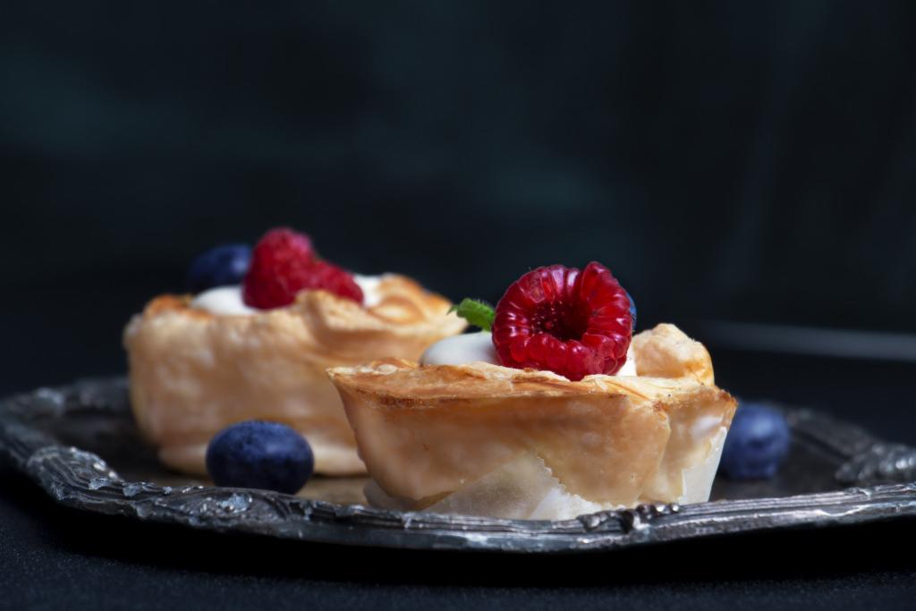 Dwa kruche babeczki z kremem, malinami i borówkami na srebrnej tacy, ciemne tło.