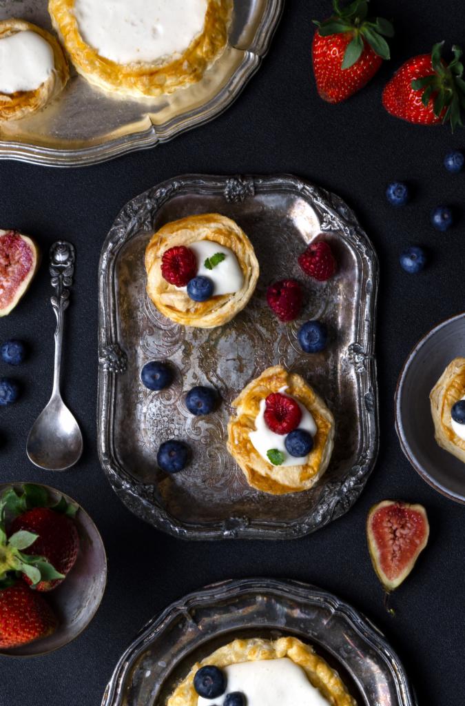 Tarty z kremem i owocami na srebrnych paterach, widok z góry, ciemne tło, figi, truskawki i łyżeczka obok.