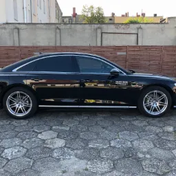 Czarna limuzyna Audi A8 z przyciemnionymi szybami bocznymi, zaparkowana na kostce brukowej, z widocznym logo firmy 'DECENT HAUSE' na drzwiach. Tło stanowi drewniany płot i budynki.