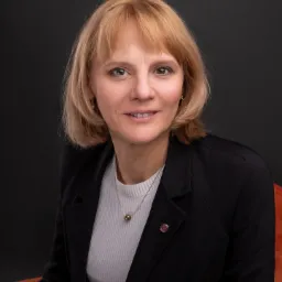 Portret uśmiechniętej kobiety w czarnej marynarce i białym golfie na ciemnoszarym tle, siedzącej na pomarańczowym krześle.