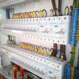 Rozdzielnica elektryczna z dwoma rzędami zabezpieczeń Hager, okablowaniem oznaczonym żółtymi numerami i narzędziami (kombinerki, szczypce) u dołu.