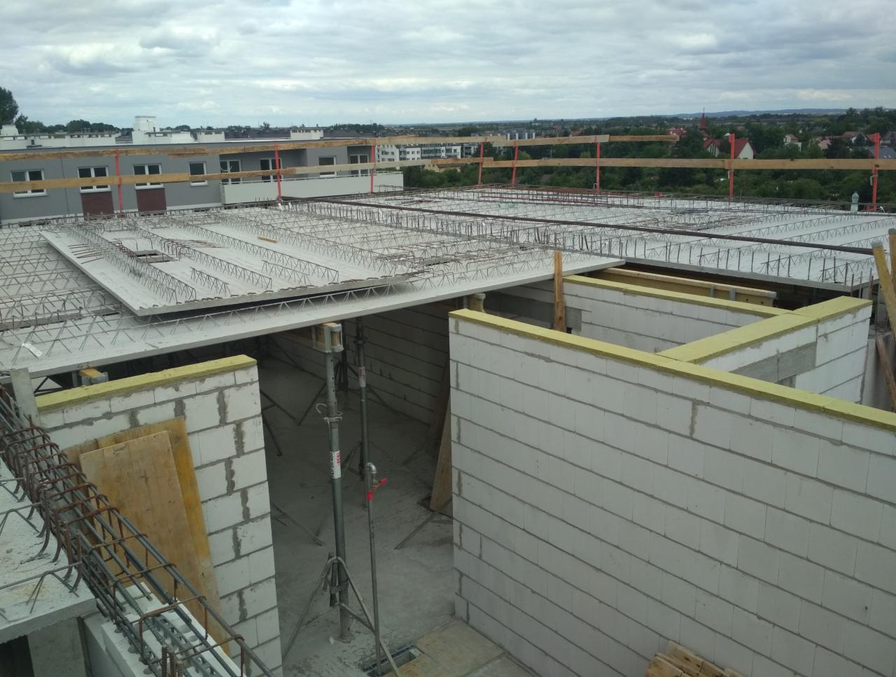 Wysoki kąt widzenia na budowę domu z betonu komórkowego, widoczny strop żelbetowy z siatką zbrojeniową, ściany z izolacją z wełny mineralnej, stalowe podpory tymczasowe i drewniane zabezpieczenia...