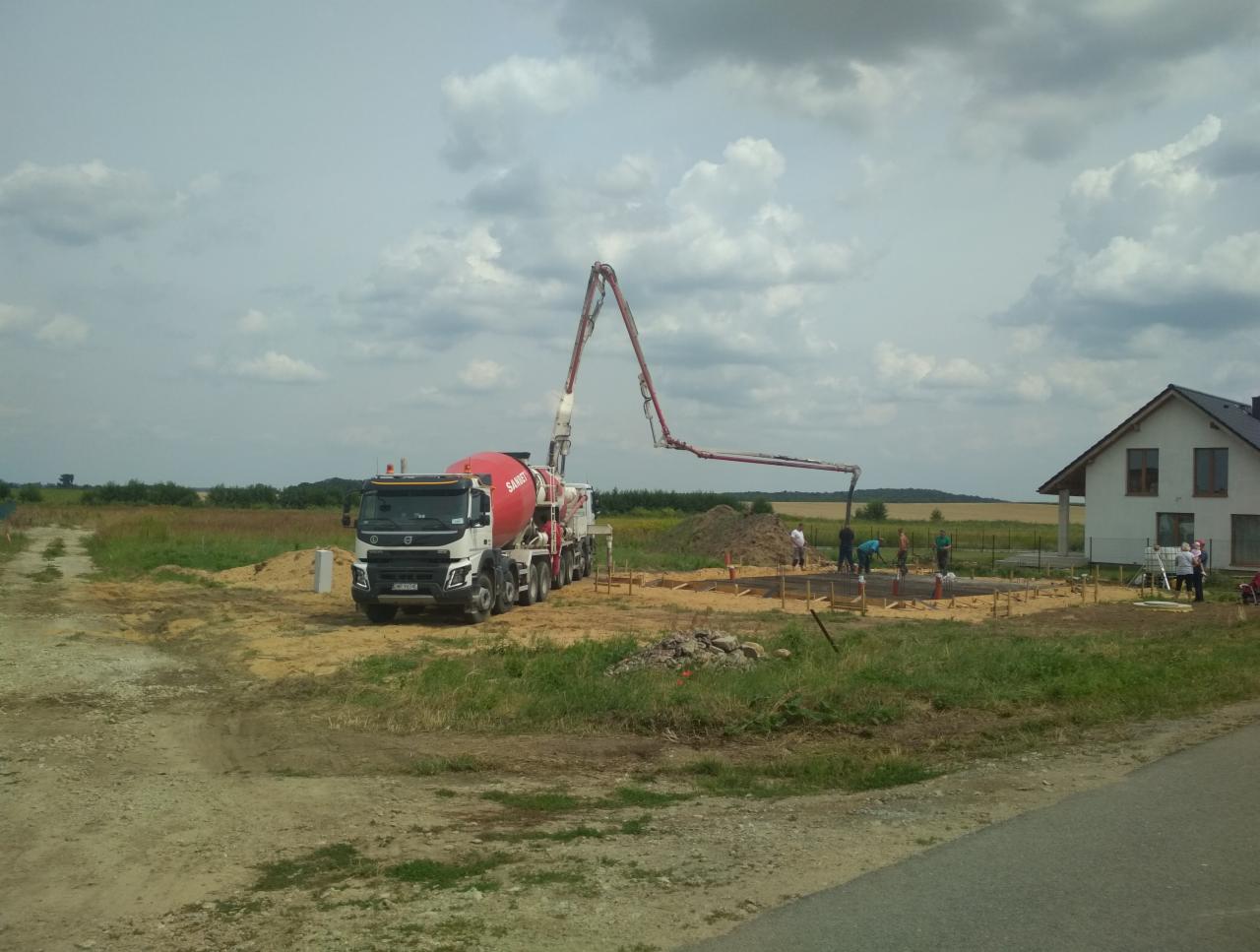 Wylewanie betonu pod fundament domu jednorodzinnego z użyciem pompy do betonu, widoczny szalunek, pracownicy budowlani i krajobraz wiejski w tle.