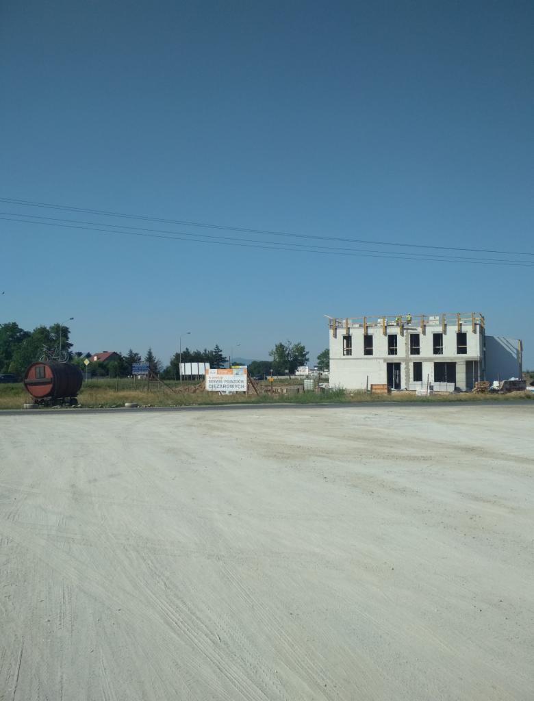 Surowy budynek żelbetowy w trakcie budowy, widoczni pracownicy na drugim piętrze, otoczony rozległym betonowym placem i zielenią w tle.