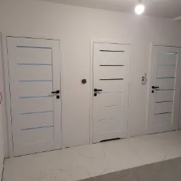 Trzy białe, nowoczesne drzwi wewnętrzne z czarnymi klamkami, z różnymi wzorami poziomych pasków, zamontowane w nowym, ale jeszcze nie wykończonym pomieszczeniu z surową podłogą i wystającą żarówką.