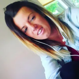 Kobieta z ombre włosami, słuchawkami i naszyjnikiem z puzzli, uśmiecha się w kierunku aparatu, pozując na tle okna i jasnej ściany.