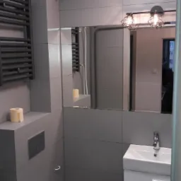 Szara łazienka z grzejnikiem drabinkowym, lustrem, umywalką i toaletą w zabudowie, widoczna przez uchylone drzwi.