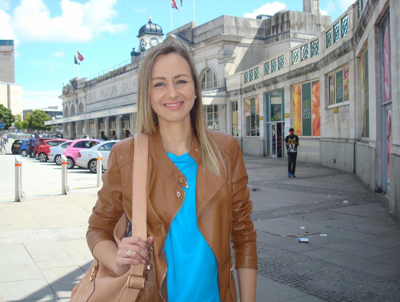 Uśmiechnięta blondynka w brązowej skórzanej kurtce na tle budynku Great Western Railway w Swindon, Anglia.