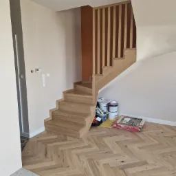 Jasne schody z drewnianą balustradą i parkietem w jodełkę, kontrastujące z szarą posadzką. Widoczne puszki z farbą i narzędzia malarskie.