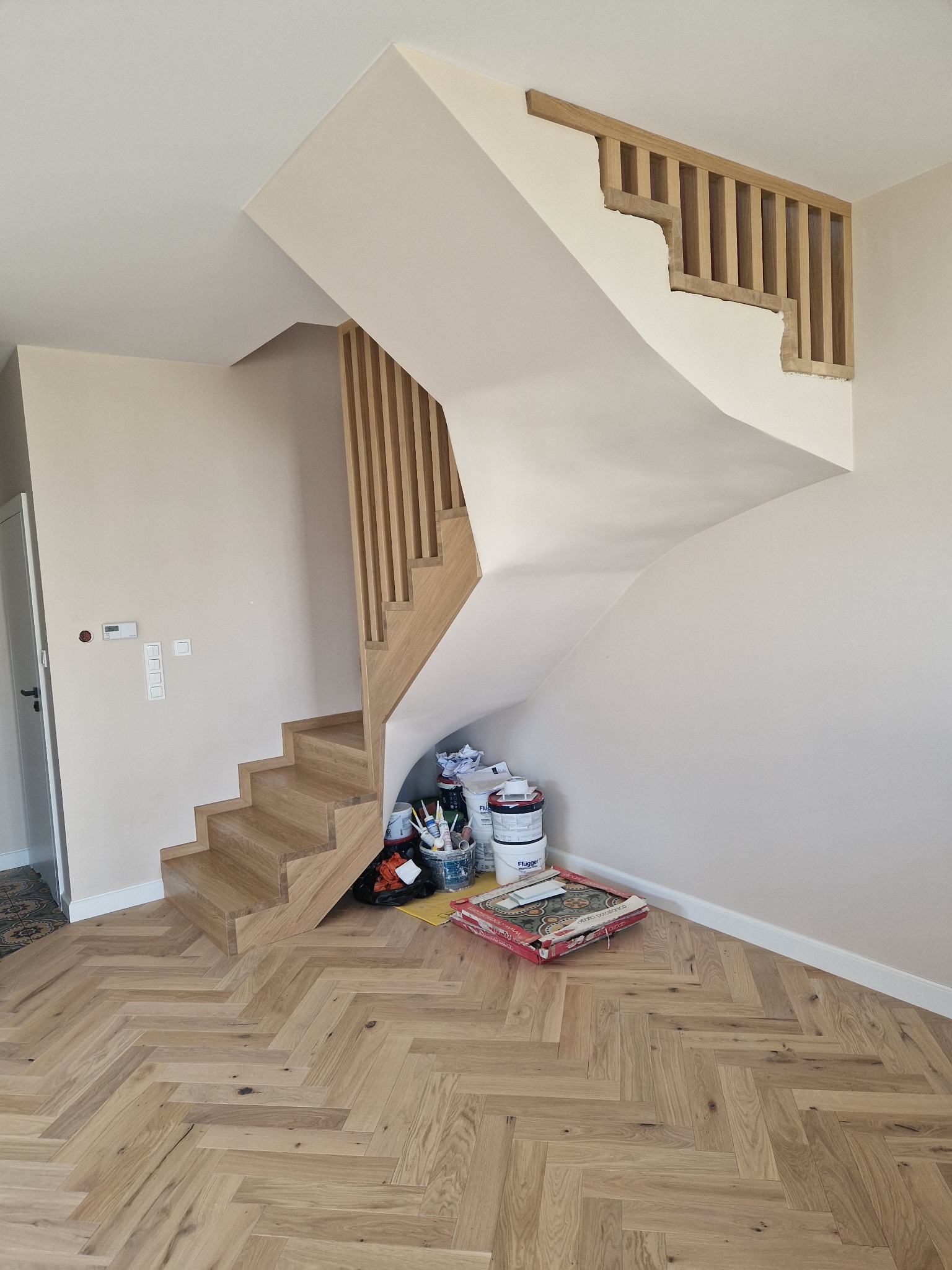 Kaskadowe schody drewniane z balustradą z pionowych szczebli, wykończone w jasnym odcieniu drewna. Parkiet w jodełkę, farby malarskie i narzędzia pod schodami.