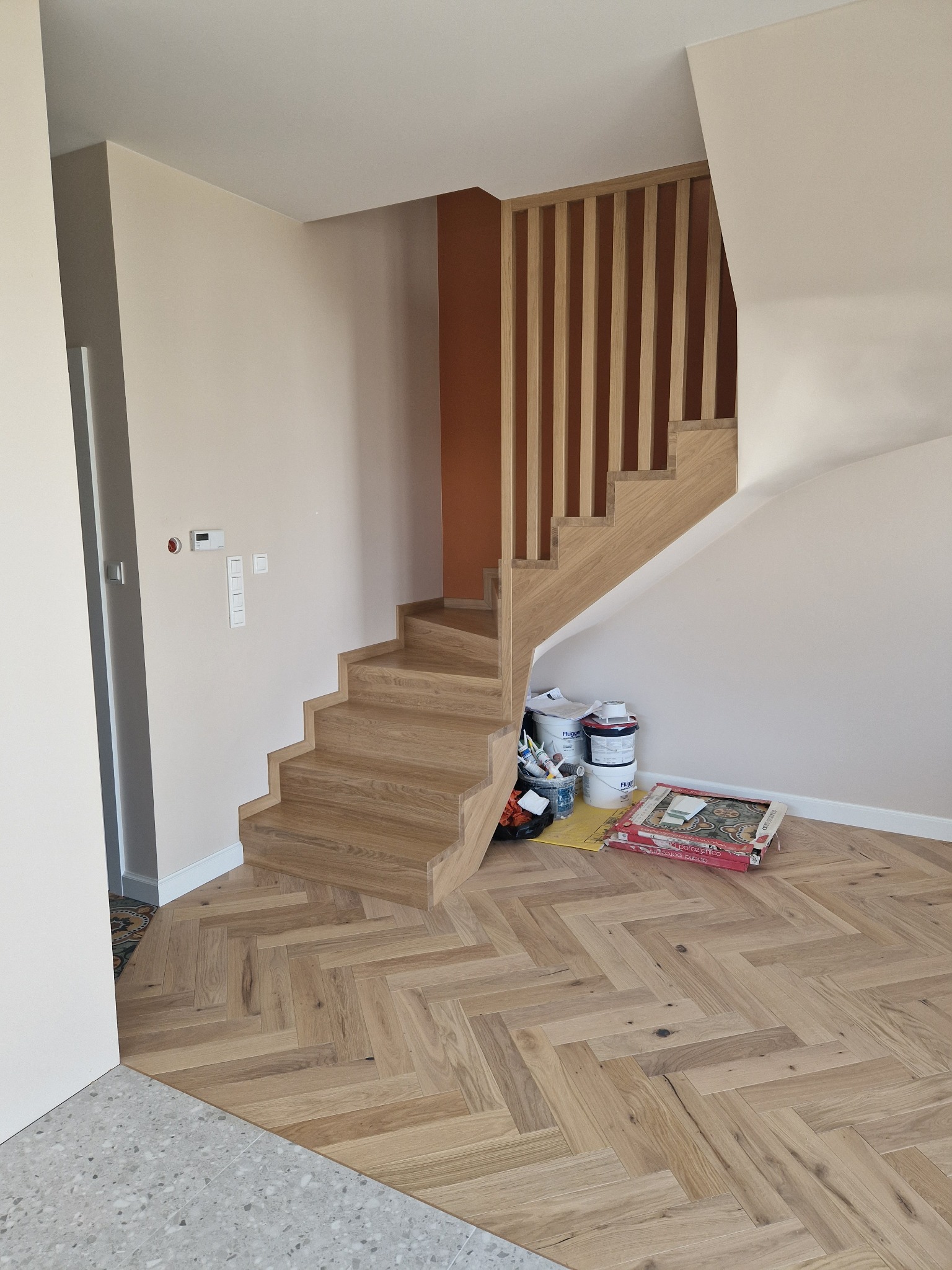 Jasne schody z drewnianą balustradą i parkietem w jodełkę, kontrastujące z szarą posadzką. Widoczne puszki z farbą i narzędzia malarskie.