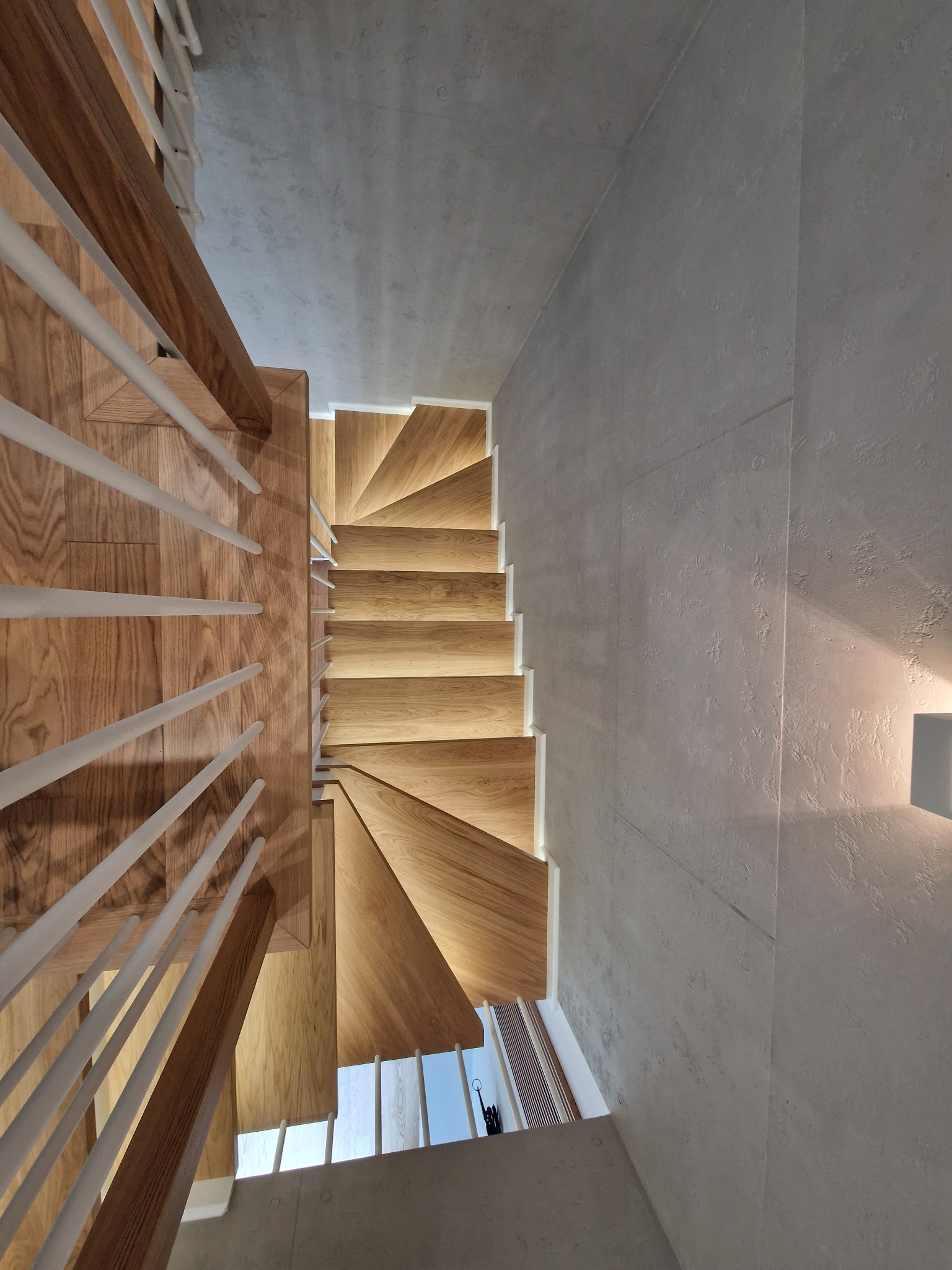 Nowoczesne schody drewniane z białymi balustradami i subtelnym podświetleniem LED, widok z góry, przylegające do betonowej ściany o minimalistycznym wykończeniu.