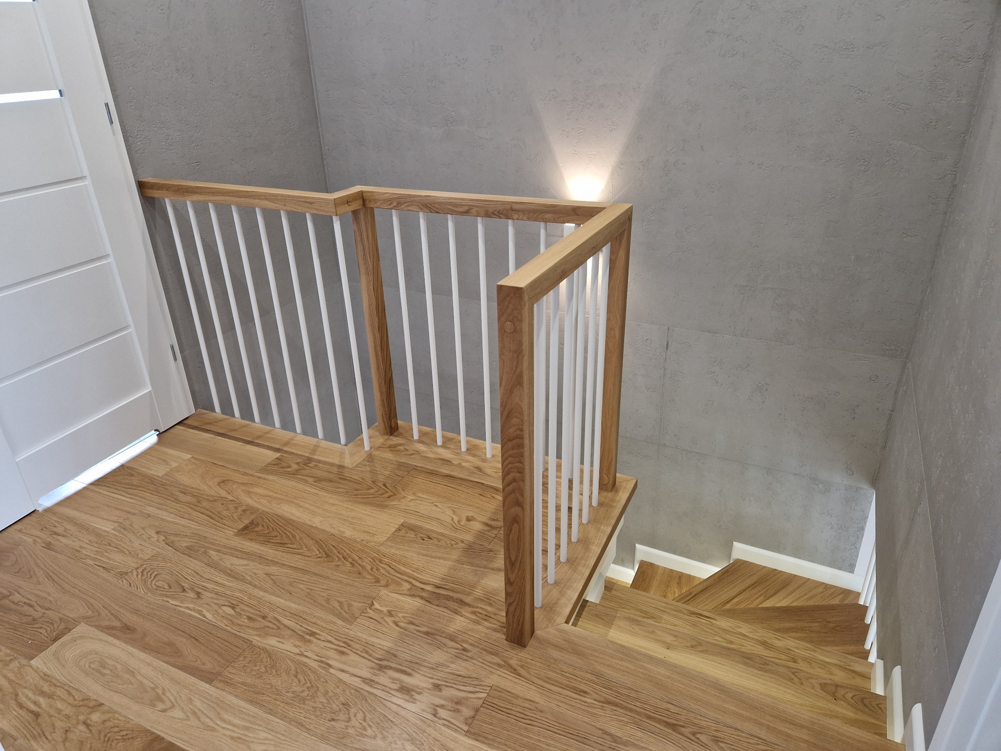 Jasne schody z drewnianymi stopniami i białymi balustradami, oświetlone punktowo, na tle szarej ściany z fakturą betonu i białych drzwi. Widok z góry.