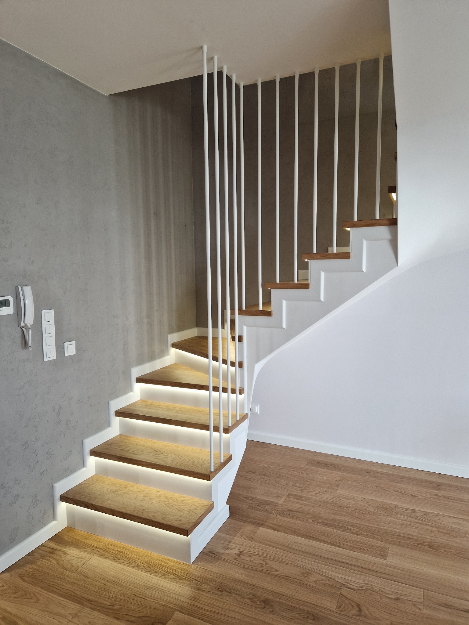 Nowoczesne schody z podświetlanymi stopniami, drewniane z białymi elementami konstrukcyjnymi i pionowymi barierkami. Szara ściana w tle, drewniana podłoga.