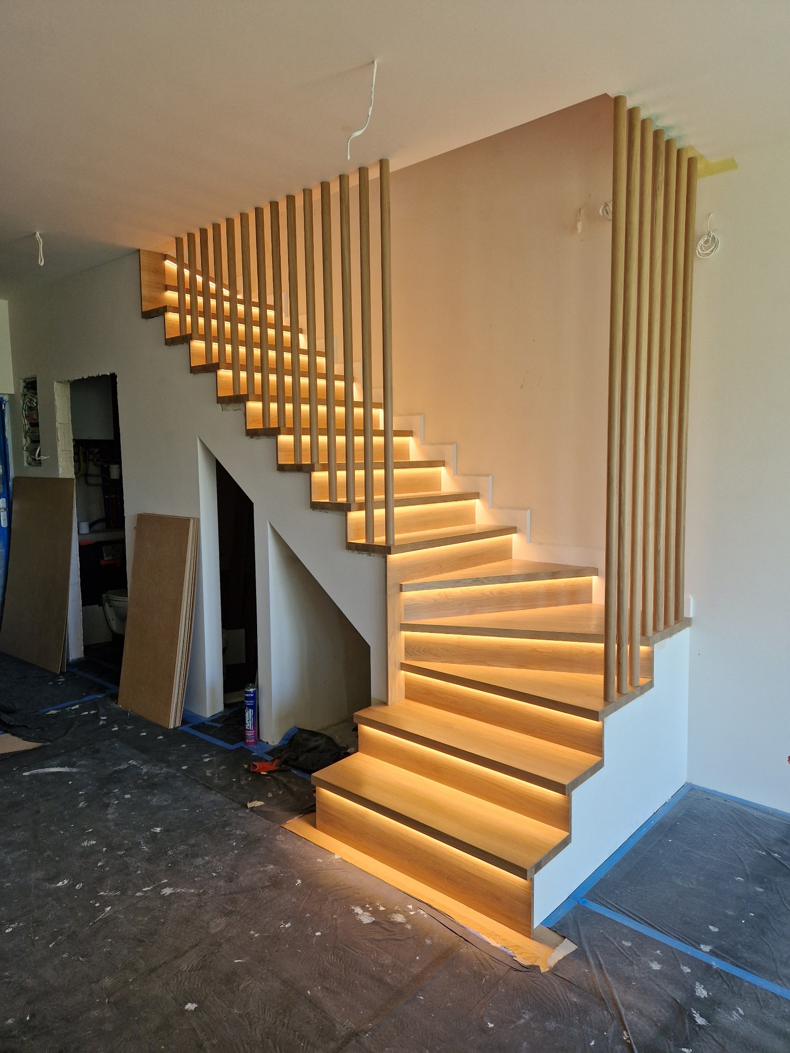 Nowoczesne schody drewniane z podświetleniem LED, zabezpieczone folią podłogi, widoczne pionowe elementy dekoracyjne.
