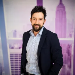 Portret uśmiechniętego mężczyzny z brodą w marynarce na tle grafiki z drapaczami chmur, sugerujący profesjonalizm w branży finansowej lub nieruchomości.