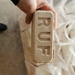 Dłoń trzyma brykiet RUF na tle worków z brykietem. Widoczne detale tekstury brykietu i wytłoczone litery RUF. Prezentacja produktu.