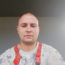 Autoportret pracownika remontowego z Ruda Śląska w ubraniu roboczym, z plamami farby. Widoczny fragment pomalowanej ściany w tle. Ujęcie od przodu.