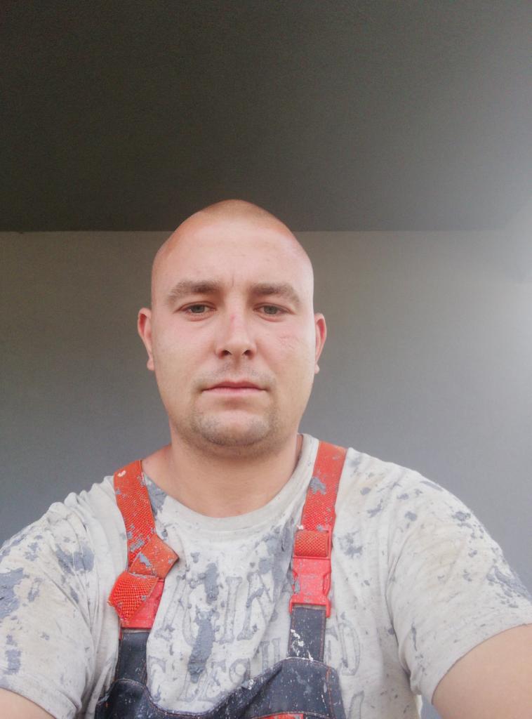 Autoportret pracownika remontowego z Ruda Śląska w ubraniu roboczym, z plamami farby. Widoczny fragment pomalowanej ściany w tle. Ujęcie od przodu.