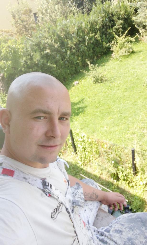 Portret uśmiechniętego mężczyzny z tatuażem na ramieniu, ubrany w roboczą koszulkę z plamami farby, na tle zieleni ogrodu.