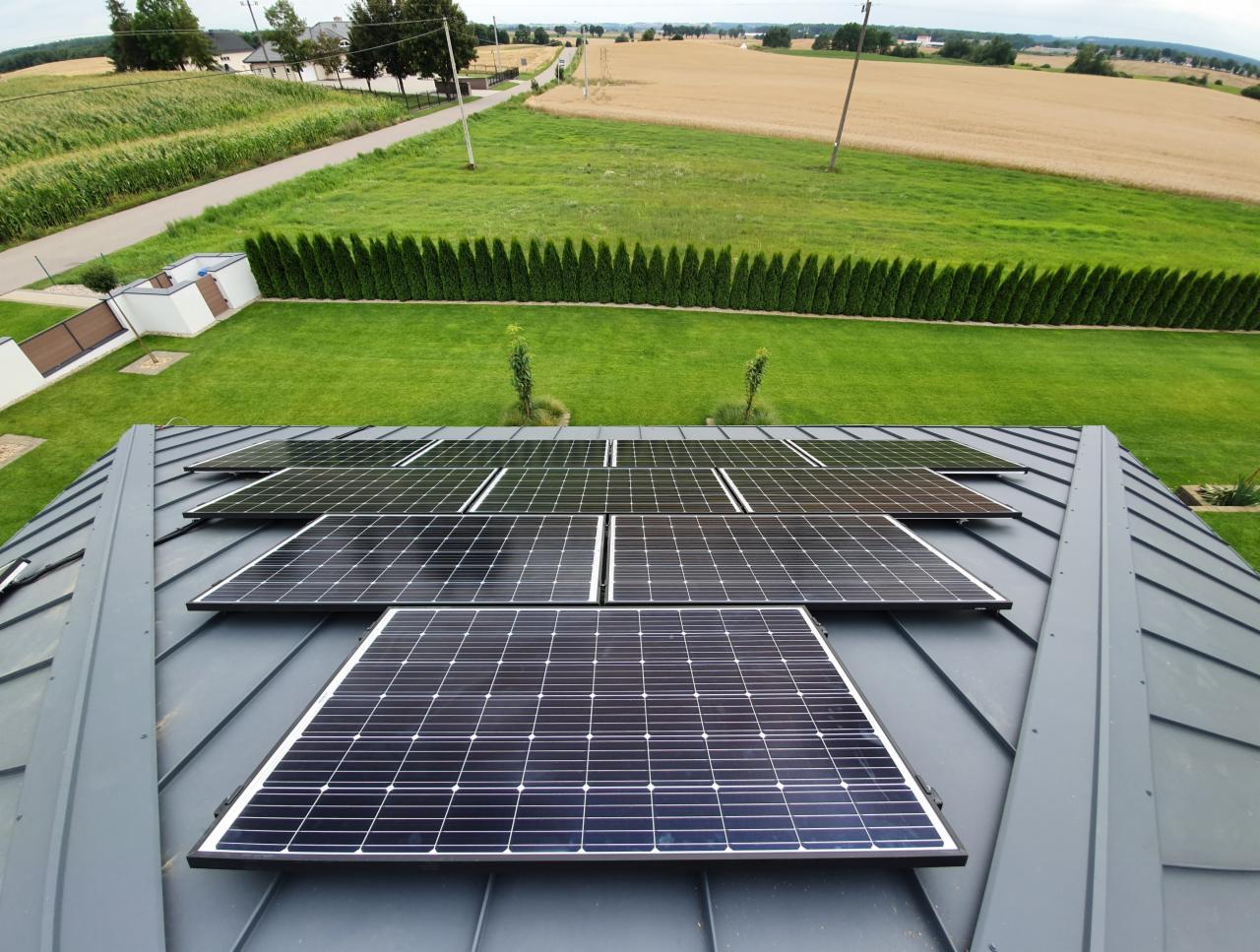 Panele fotowoltaiczne zamontowane na szarym, metalowym dachu domu jednorodzinnego z widokiem na zadbany trawnik, rząd tuj i pola uprawne w tle. Perspektywa z góry.