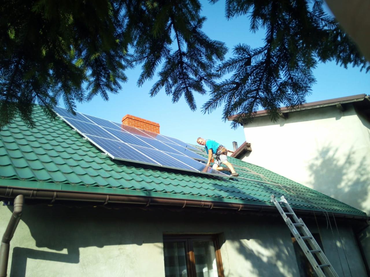 Instalator w niebieskiej koszulce montuje panele na zielonym dachu domu jednorodzinnego z ceglanym kominem, drabina oparta o ścianę.