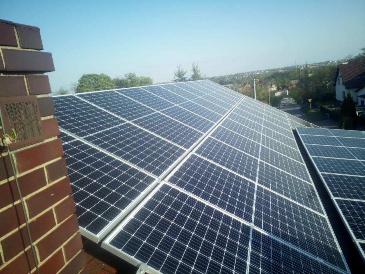 Panele słoneczne na dachu domu, widok z góry, z ceglanym kominem po lewej stronie i zabudową w tle.