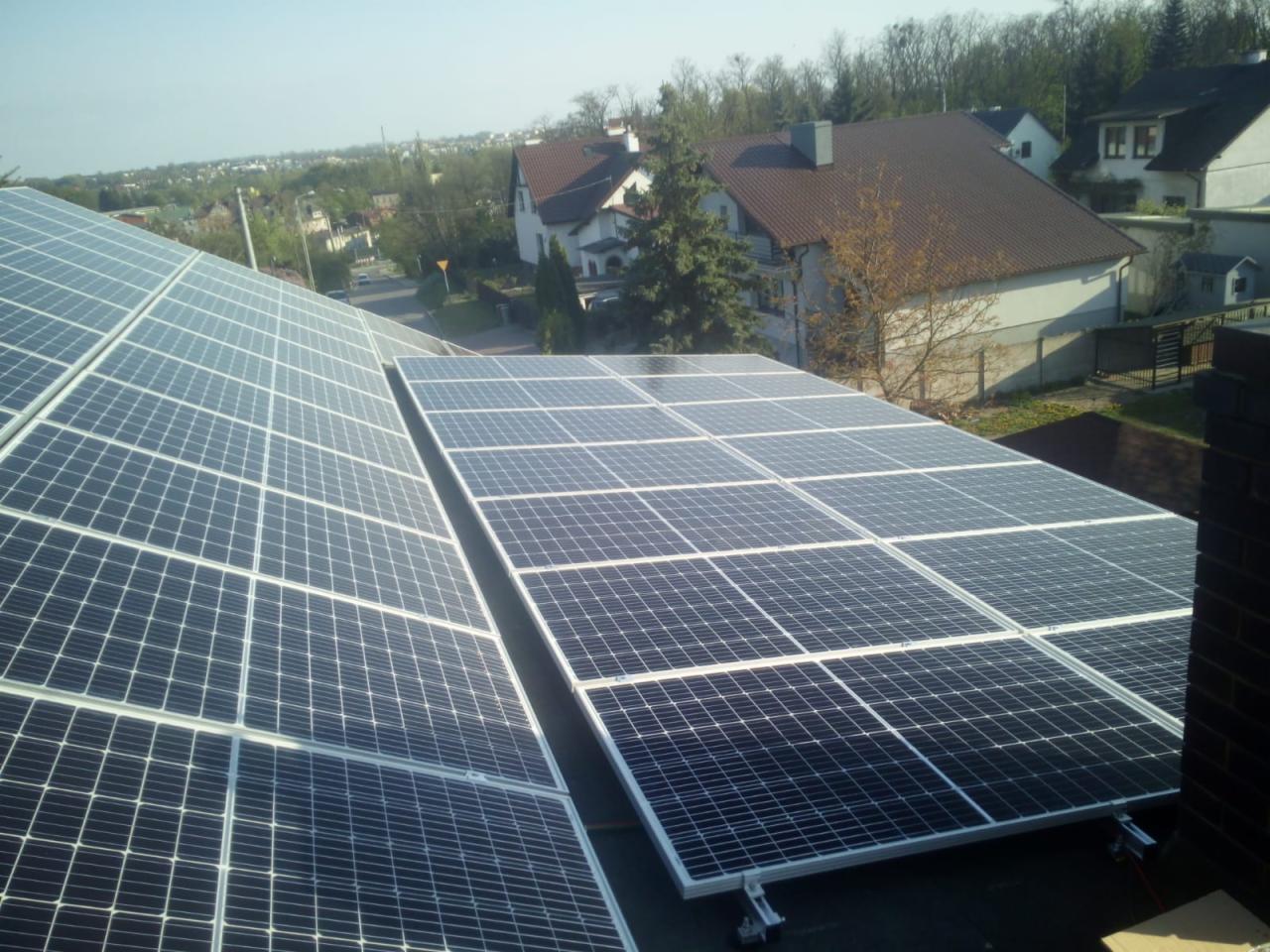 Instalacja fotowoltaiczna na dachu domu jednorodzinnego, widok z góry na panele słoneczne w regularnym układzie, w tle zabudowa mieszkalna i drzewa.