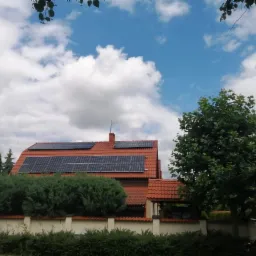 Dom jednorodzinny z zainstalowanymi panelami słonecznymi na czerwonym dachu, widok z poziomu ulicy w słoneczny dzień.