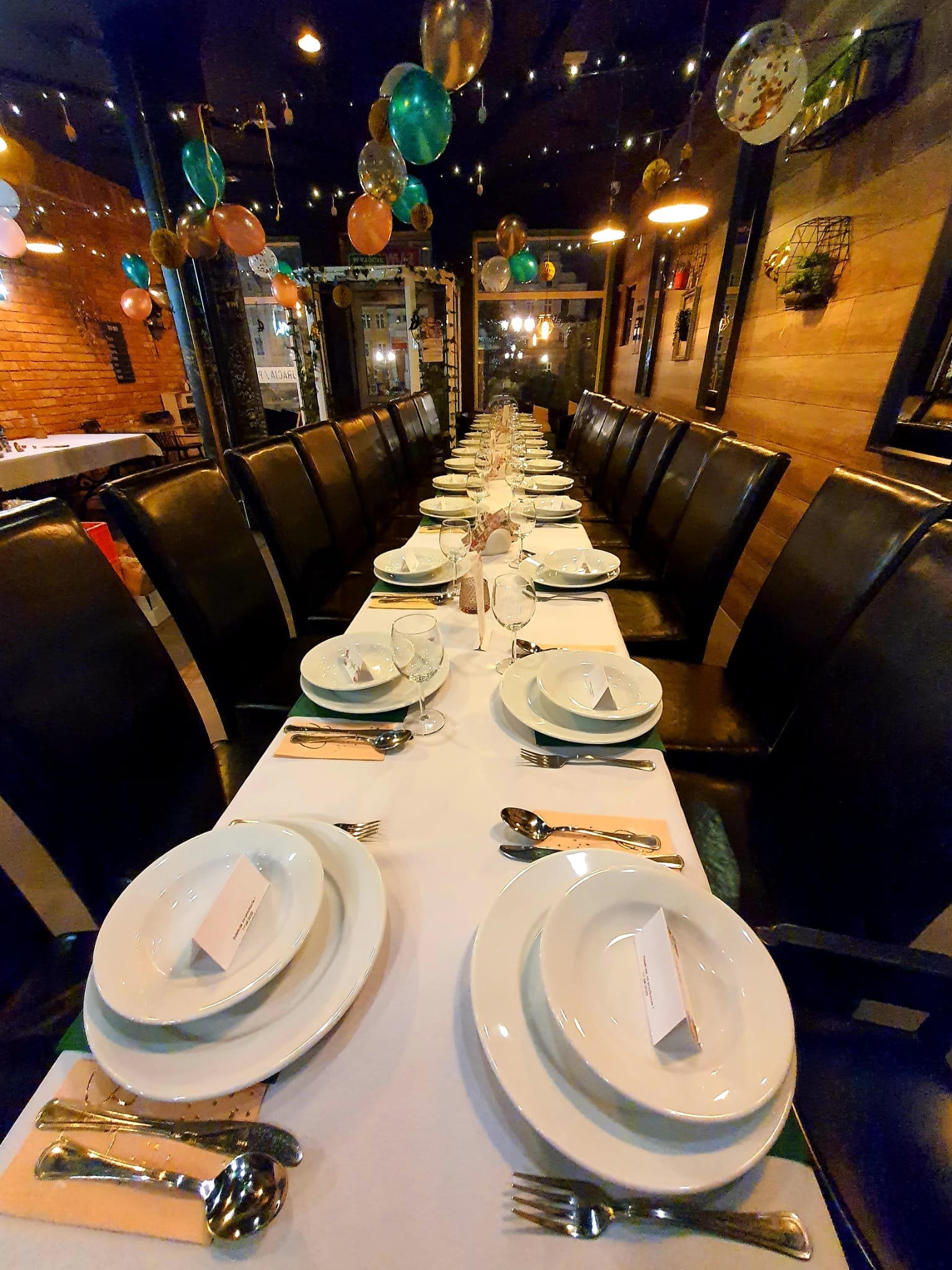Długi stół nakryty białym obrusem, z zastawą dla wielu gości, udekorowany balonami pod sufitem, ustawiony w restauracji o ceglanych ścianach.