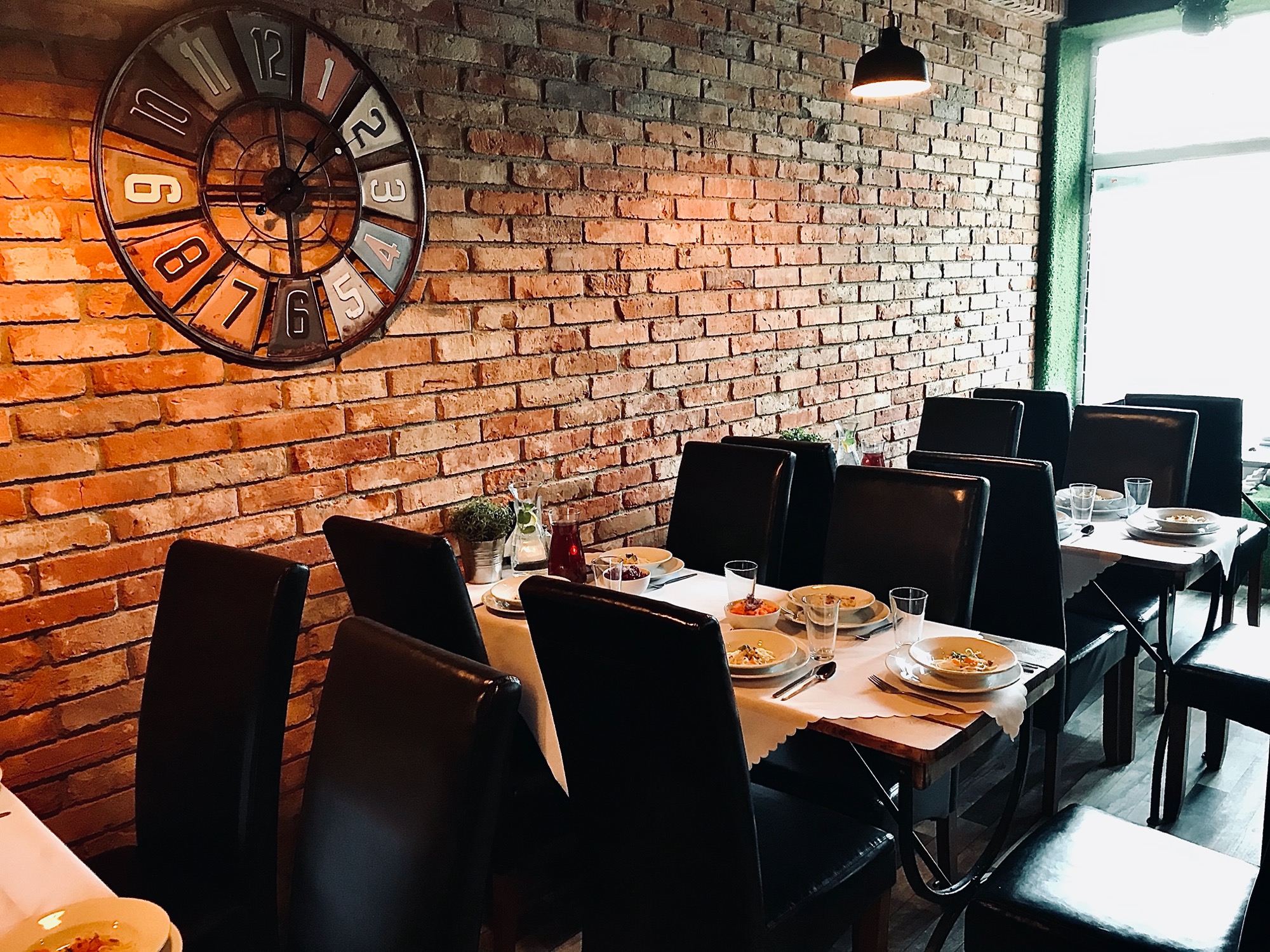Ustawione stoły z białymi obrusami i nakryciami, gotowe na przyjęcie gości w rustykalnym wnętrzu z ceglaną ścianą i dekoracyjnym zegarem.