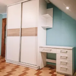 Biała szafa przesuwna z drewnianymi akcentami, biurko z szufladami i półki w pokoju na poddaszu z parkietem w jodełkę.