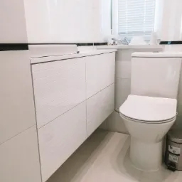 Biała łazienka z wiszącą szafką pod umywalką z poziomymi frezowaniami, toaletą i koszem na śmieci z metalu.