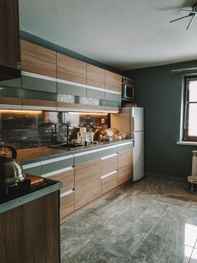 Nowoczesna kuchnia z drewnianymi frontami szafek, szarymi akcentami i marmurową podłogą. Na blacie widoczne naczynia i czajnik na płycie grzewczej.