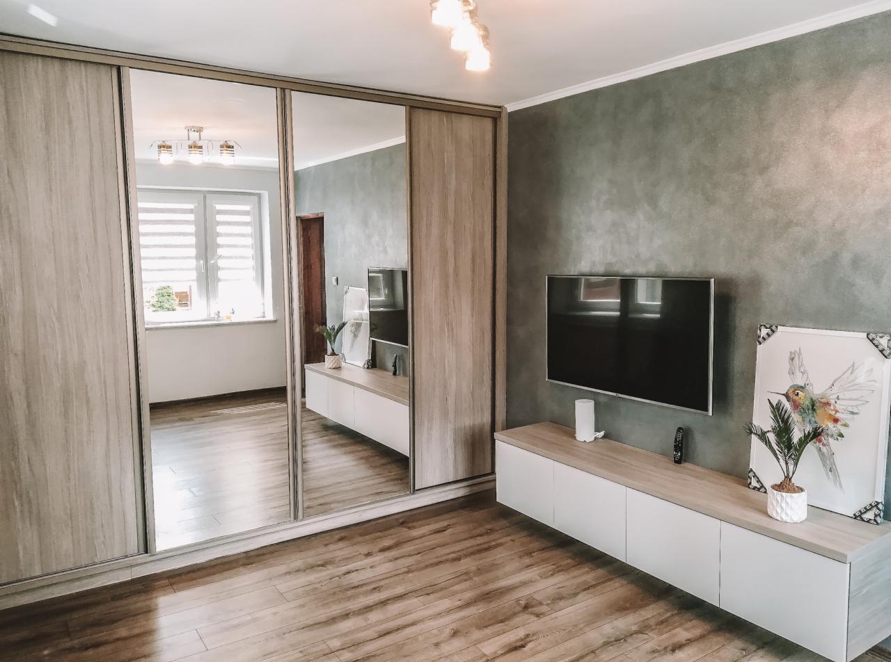 Nowoczesna szafa wnękowa z lustrzanymi drzwiami przesuwnymi, odbijająca wnętrze pokoju z oknem i białą komodą pod telewizorem, na tle szarej ściany.