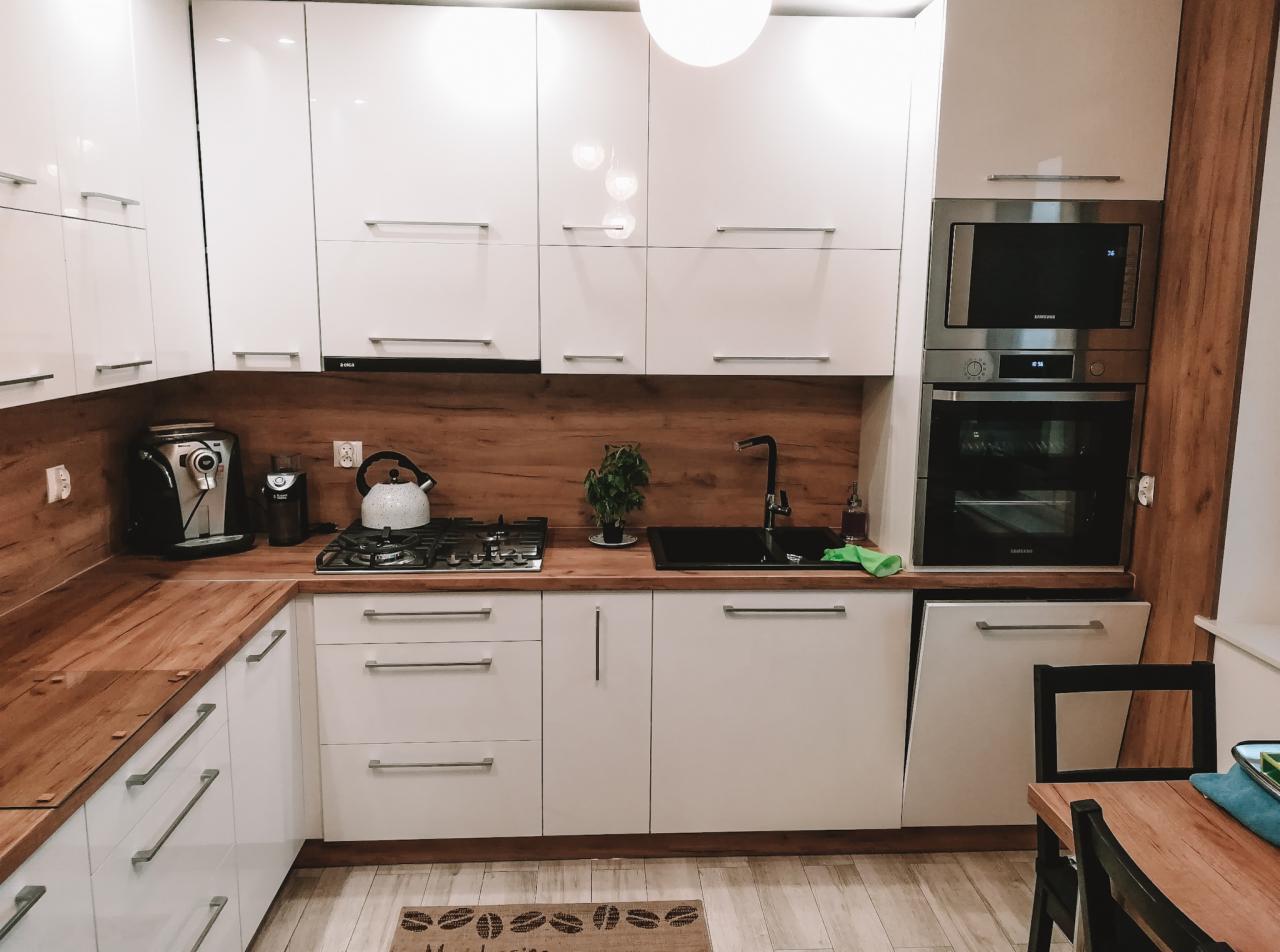 Biała kuchnia z drewnianym blatem i panelem ściennym, czarny zlew i armatura, widoczny ekspres do kawy, czajnik i roślina doniczkowa, dywanik z motywem ziaren kawy.