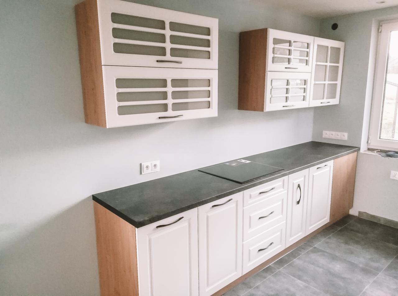 Nowoczesna kuchnia z białymi szafkami dolnymi i górnymi z drewnianymi bokami, szarym blatem i płytą indukcyjną, widok z boku na ścianę z gniazdkami.