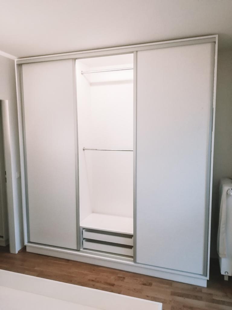 Biała szafa przesuwna z otwartą środkową częścią, widoczne półki i dwie szuflady, minimalistyczny design.