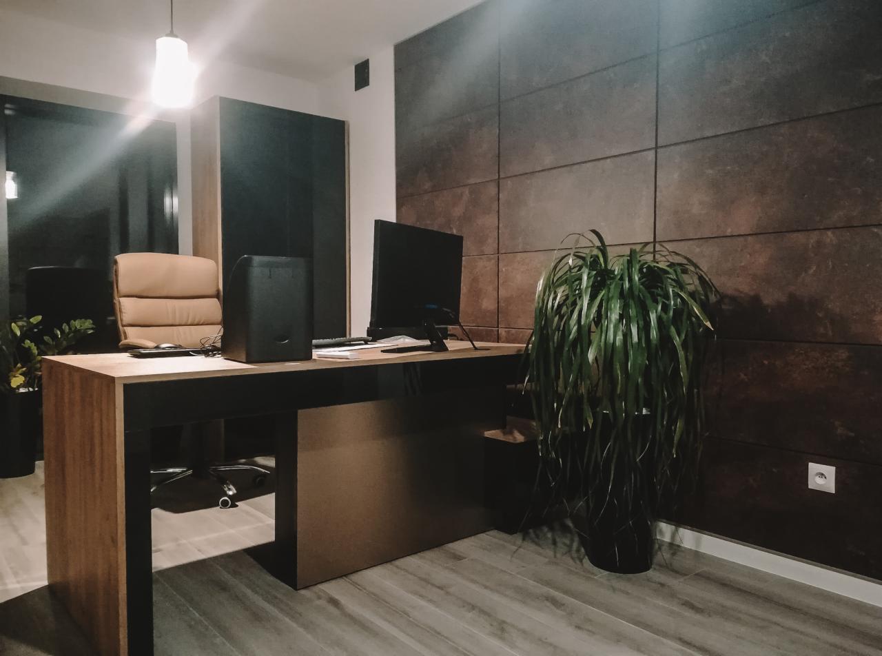 Nowoczesne biuro z drewnianym biurkiem, ergonomicznym fotelem, dużym monitorem i ścianą z paneli dekoracyjnych w odcieniach brązu, oświetlone wiszącą lampą.
