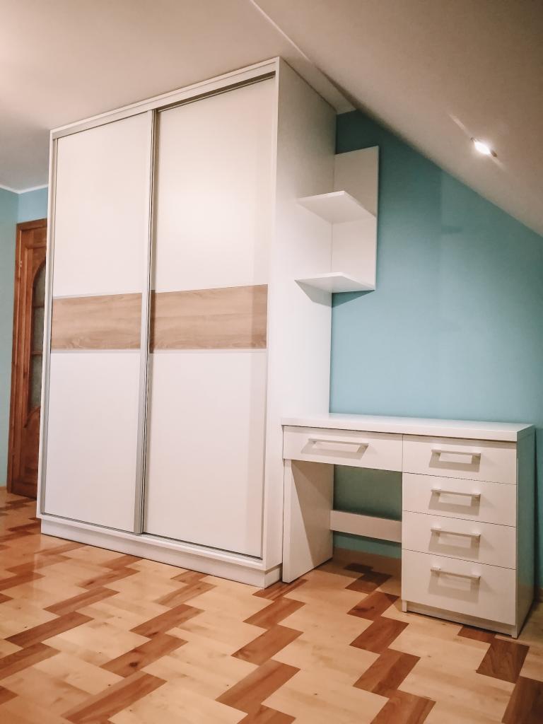 Biała szafa przesuwna z drewnianymi akcentami, biurko z szufladami i półki w pokoju na poddaszu z parkietem w jodełkę.