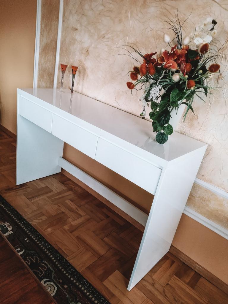 Biała, lakierowana konsola z trzema szufladami, ustawiona w przedpokoju z parkietem w jodełkę i dekoracyjną ścianą, ozdobiona bukietem kwiatów i dwoma kieliszkami.