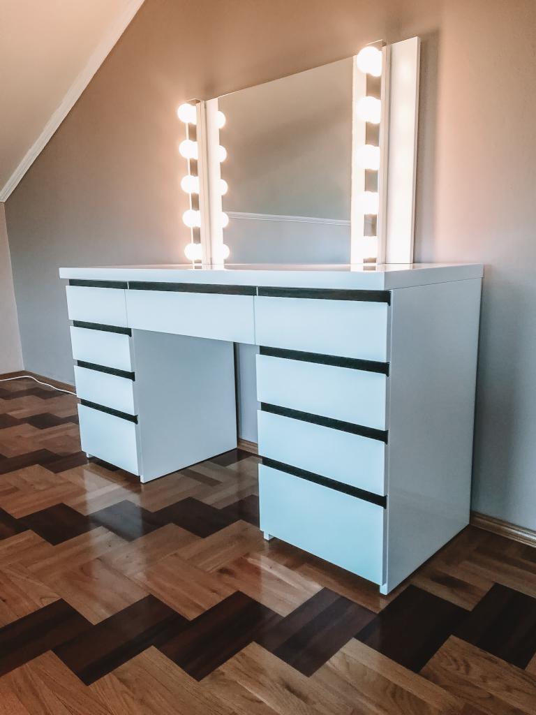 Biała toaletka z lustrem podświetlanym żarówkami, z szufladami z czarnymi uchwytami, ustawiona na parkiecie w jodełkę, w pokoju na poddaszu.