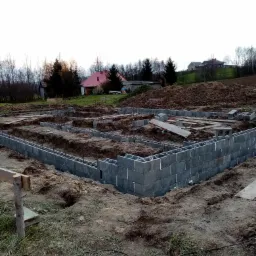Wznoszenie fundamentów z bloczków betonowych na terenie budowy domu jednorodzinnego, widoczne zbrojenia i przygotowane wykopy, w tle zabudowa wiejska.