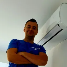 Uśmiechnięty mężczyzna w niebieskiej koszulce z napisem 'KLIMATYZACJE' stoi pod zamontowanym, białym klimatyzatorem na ścianie.