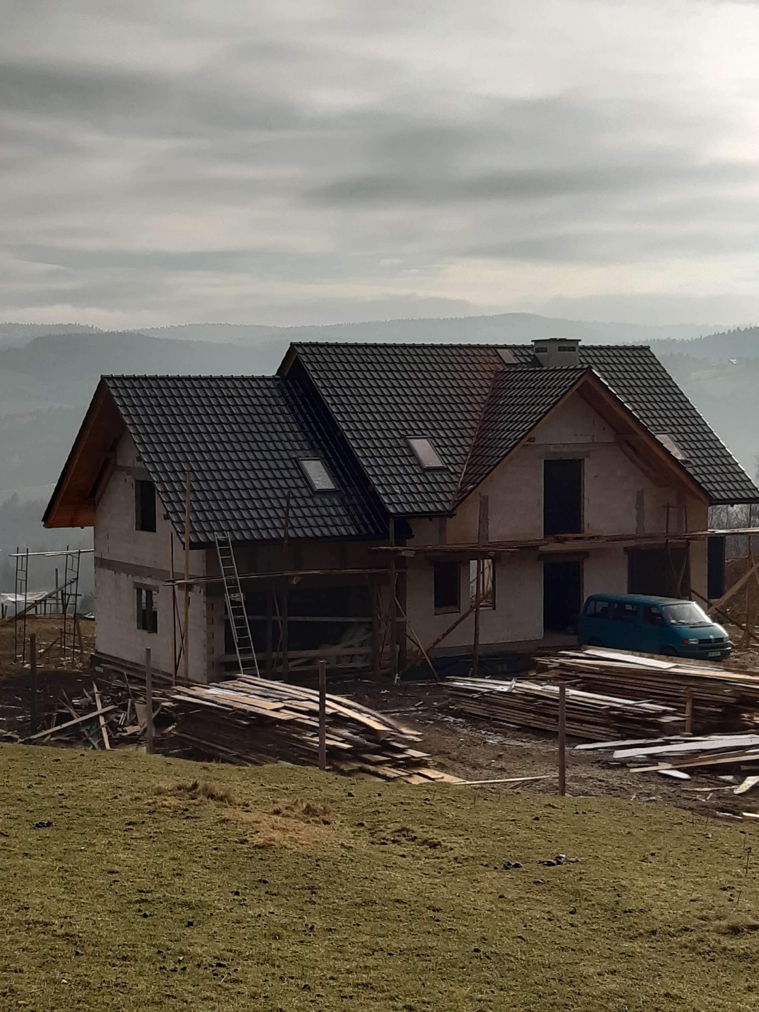 Niedokończony dom murowany z ciemną dachówką i oknami dachowymi, otoczony rusztowaniami i stertą drewna, na tle zalesionych wzgórz w pochmurny dzień.
