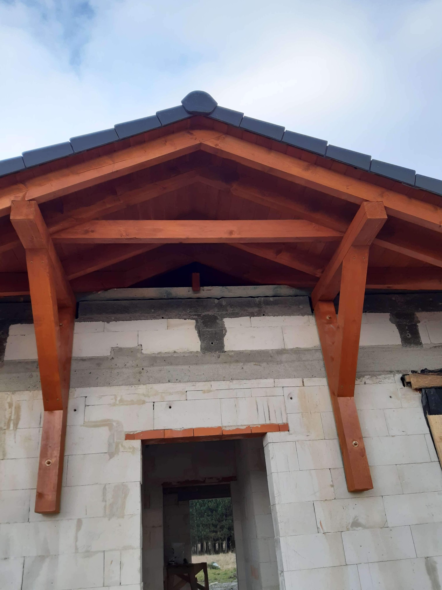 Surowy stan domu z bloczków, widoczna drewniana konstrukcja dachu i fragment pokrycia dachowego z ciemnej dachówki, otwór drzwiowy.