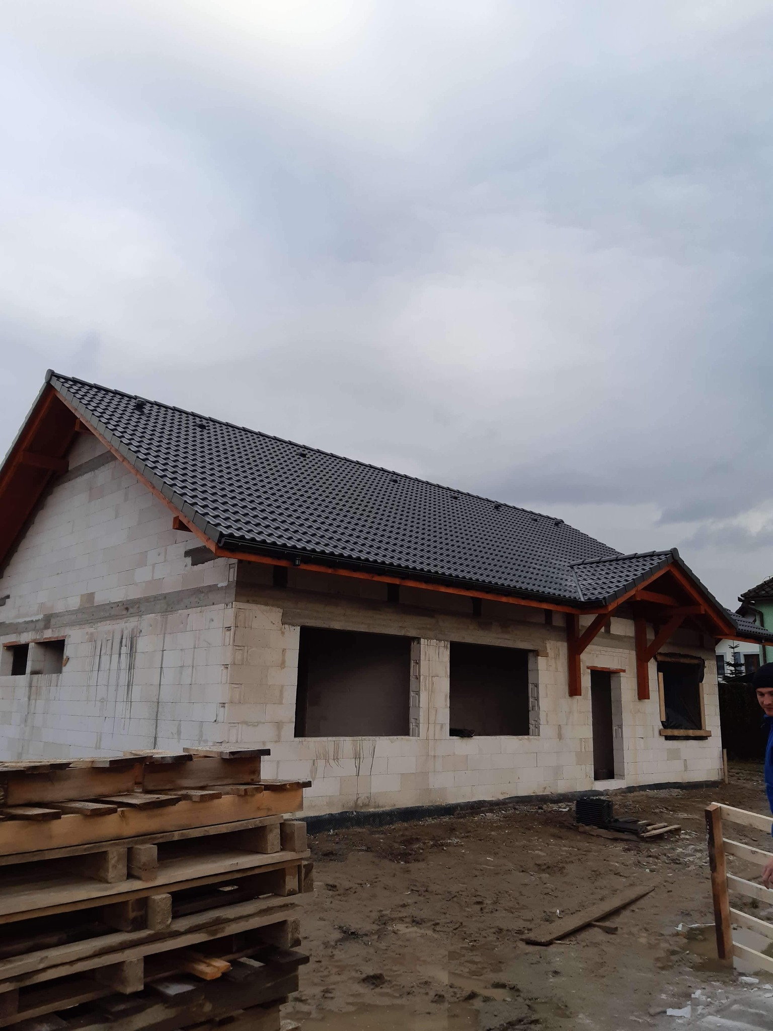 Surowy stan domu z białego bloczka z czarnym dachem, widoczne drewniane elementy konstrukcyjne, palety z materiałami budowlanymi na pierwszym planie, pochmurne niebo w tle.