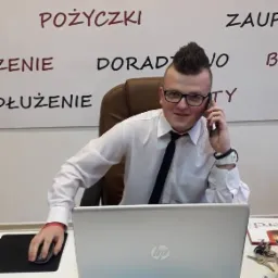 Młody doradca finansowy w okularach, rozmawia przez telefon przy laptopie. W tle ściana z napisem 'Pożyczki', 'Doradztwo'.