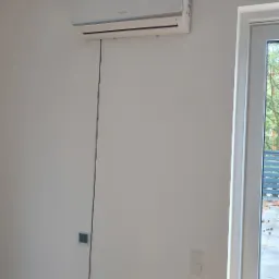 Biała klimatyzacja zamontowana na ścianie w jasnym pokoju, widoczny kabel zasilający biegnący wzdłuż ściany, okno z drewnianym parapetem po prawej stronie.