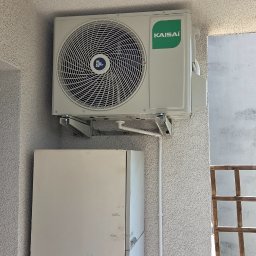 INSTALSERW - ROMAN RACZEK - Biały agregat klimatyzacji Kaisai z zielonym logo, zamontowany na wspornikach do jasnej, chropowatej ściany zewnętrznej, obok białej szafki. Widoczne rurki instalacyjne.